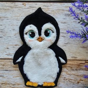 Könnte beinhalten: Ein gestickter Plüschaufnäher in Form eines Pinguinbabys mit schwarz-weißem Fell, großen hellblauen Augen, einem kleinen orangefarbenen Schnabel und orangefarbenen Füßen. Das Design zeichnet sich durch weiche Texturen und detaillierte Nähte aus.