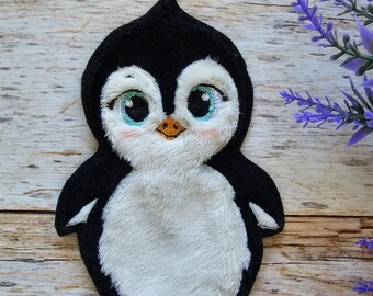 May include: A plush embroidered patch shaped like a baby penguin with black and white fur, large light blue eyes, a small orange beak, and orange feet. The design features soft textures and detailed stitching, creating a rounded, cartoon-like appearance.