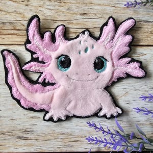 Könnte beinhalten: Ein rosa Axolotl-Aufnäher mit schwarzem Rand. Der gestickte Axolotl hat große, blau-grüne Augen und einen lächelnden Mund. Der Aufnäher befindet sich auf einer Holzoberfläche mit Lavendelzweigen.