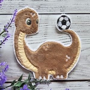 Könnte beinhalten: Ein brauner Dinosaurier-Aufnäher mit langem Hals und hellgelbem Bauch. Der Dinosaurier hat ein grünes Auge und balanciert einen schwarz-weißen Fußball auf der Spitze seines geschwungenen Schwanzes. Das Motiv ist mit weißem Garn umrandet.