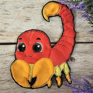 Könnte beinhalten: Ein roter und gelber Filzscorpion mit großen, schwarzen Augen. Der Skorpion hat einen eingerollten Schwanz und lächelt.