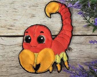 May include: A red and yellow felt scorpion with large, black eyes. The scorpion has a curled tail and is smiling.
