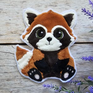 Könnte beinhalten: Ein Plüsch-Rotpanda-Applikationen mit weißen Fell-Akzenten. Der Panda hat schwarze Augen, eine schwarze Nase und einen schwarzen Mund. Die Applikation ist perfekt, um jedem Projekt einen Hauch von Verspieltheit zu verleihen.