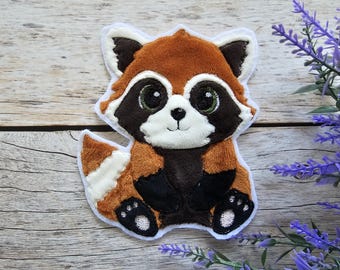 May include: An embroidered patch featuring a stylized red panda with large dark eyes, a white face mask, and a bushy tail. The design uses shades of brown, black, and white thread with a white border, depicting the animal in a seated position.