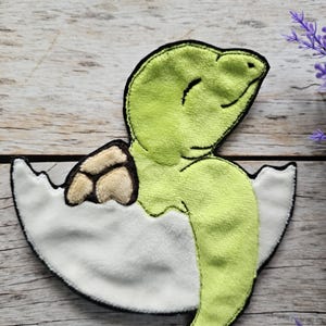 Op de afbeelding: Een pluche applicatie van een babyschildpad die uit een ei komt. De schildpad is lichtgroen met een zwarte omtrek, en het ei is wit met een bruin gedeelte. De applicatie is ontworpen om op stof te worden genaaid.