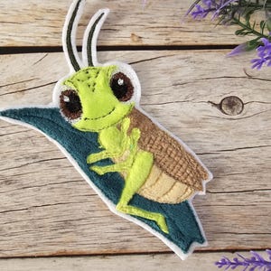 Könnte beinhalten: Ein gestickter Aufnäher mit einer Cartoon-Heuschrecke auf einem blauen Blatt. Die Heuschrecke ist grün und braun mit großen, schwarzen Augen und einem weißen Streifen auf dem Rücken.
