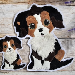 Könnte beinhalten: Zwei gestickte Filz-Hundepatchs. Der größere Patch ist ein brauner, schwarzer und weißer Hund mit einer weißen, flauschigen Brust. Der kleinere Patch ist ein brauner, schwarzer und weißer Hund mit einer weißen, flauschigen Brust. Beide Patches haben weiße Umrisse.