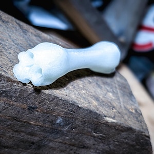 May include: A light blue, bone-shaped object with a textured surface, resting on a dark, rough wooden surface. The object has a detailed, flower-like design on one end. The lighting creates shadows, highlighting the object's form.