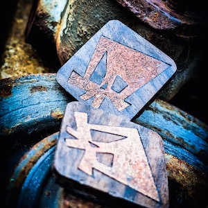 Puede incluir: Dos colgantes de madera en forma de diamante con incrustaciones de color cobre. Los colgantes presentan un diseño geométrico y están colocados sobre un fondo de metal oxidado y pintura azul.