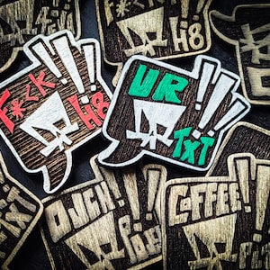 以下が含まれることがあります： 木製の吹き出し型のピン。彫刻されたテキストと感嘆符が特徴です。1つのピンには赤と白で「F*ck H8」と書かれています。別のピンには緑と白で「UR Txt」と書かれています。他のピンも同様のデザインです。