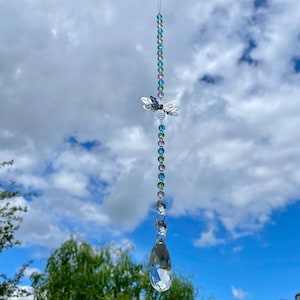 Puede incluir: Un atrapasoles decorativo con un acento metálico en forma de abeja. El atrapasoles presenta una cadena de cuentas coloridas y un colgante de cristal grande en forma de lágrima. El fondo es un cielo azul brillante con nubes blancas esponjosas.