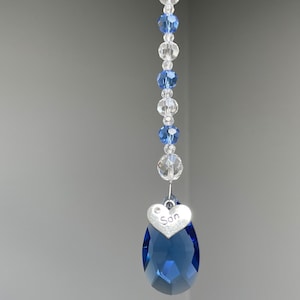 May include: A crystal suncatcher with a blue teardrop crystal and a silver heart charm with the word "Son" inscribed on it. The suncatcher is strung with clear and blue beads.