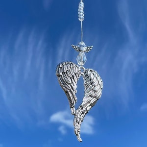 Puede incluir: Alas de ángel plateadas en forma de corazón colgando de una cadena de cuentas de cristal contra un cielo azul.