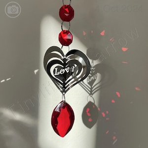 Op de afbeelding: Een zilveren hartvormige windgong met het woord "Love" erop. De gong hangt aan een touwtje met rode kristallen kralen.