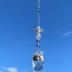 May include: A crystal suncatcher with a silver angel charm and a heart-shaped charm with the word "Son" inscribed. The suncatcher is hanging from a blue beaded string against a blue sky.