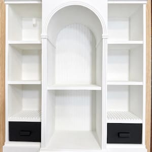 Puede incluir: Una estantería de pared blanca con una sección central arqueada con fondo de paneles verticales, flanqueada por dos secciones laterales con estantes abiertos. Cada sección lateral incluye un cajón negro en la base con un tirador rectangular.