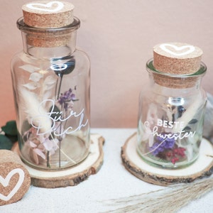 Op de afbeelding: Twee glazen potten met kurken deksels en witte harten erop. De potten zijn gevuld met gedroogde bloemen en hebben handgeschreven berichten erop. De grotere pot zegt "Für Dich" en de kleinere pot zegt "Beste Schwester".