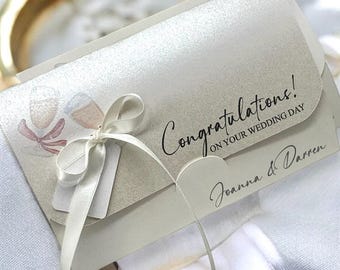 May include: A wedding invitation with a champagne glass illustration and the words "Congratulations! ON YOUR WEDDING DAY". The invitation is tied with a white ribbon and includes the names "Joanna & Darren".