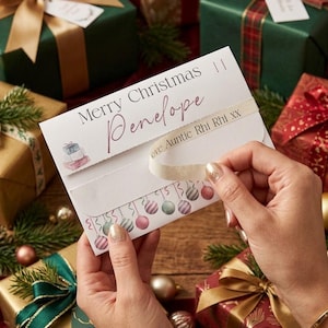 May include: A white rectangular Christmas card with the text Merry Christmas and the name Penelope in script. The card features a small illustration of stacked gifts and a border of hanging ornaments, with a cream ribbon reading Love Auntie Rhi Rhi XX.
