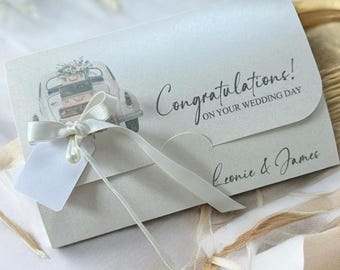 May include: A light gray wedding card with a vintage car illustration carrying flowers. The card reads "Congratulations! On your wedding day" and is tied with a white ribbon and tag. The names "Leonie & James" are printed on the card.