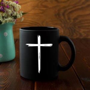 May include: A black ceramic mug with a white distressed cross design. The mug is on a wooden surface.