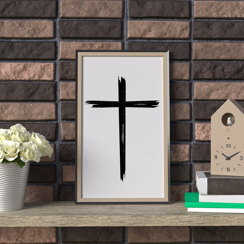 May include: A black and white framed print of a simple cross design on a white background. The print is hanging on a brick wall.