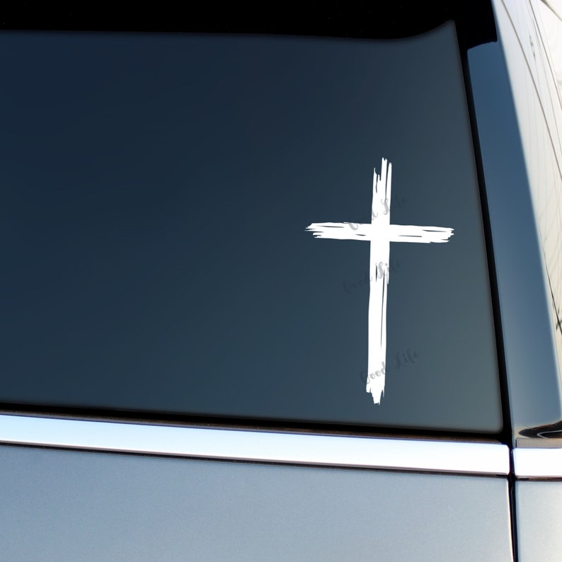 May include: White vinyl decal of a cross with a distressed, brushstroke design. The cross is on a dark gray car window.