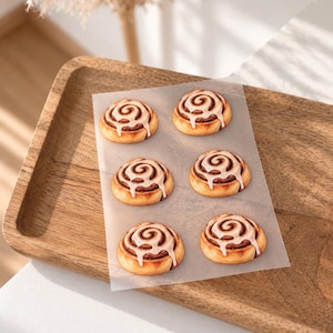 Könnte beinhalten: Sechs goldbraune Zimtschnecken, angeordnet in zwei Spalten auf einem Blatt durchscheinendem Backpapier. Jedes spiralförmige Gebäckstück zeigt eine deutliche Zimtfüllung und ist mit einem Streifen weißer Glasur verziert.