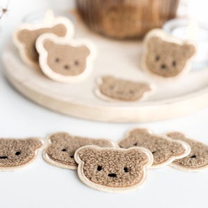 Può includere: Una collezione di toppe a forma di orsetto marrone con bordo color crema. Le toppe hanno un aspetto strutturato e sono disposte su un vassoio di legno e su una superficie bianca. Gli orsetti hanno piccoli occhi neri e una bocca cucita.