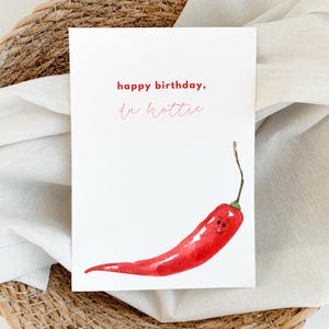 Può includere: Un biglietto bianco con il testo "happy birthday, you hottie" in rosso e rosa. Un peperoncino rosso con un viso sorridente è illustrato sotto il testo.