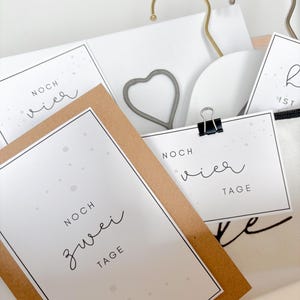May include: A close-up of a gift box containing white cards, a heart-shaped metal clip, and gold and silver hangers. The cards are printed with the words "NOCH zwei TAGE" and "NOCH vier TAGE" in a handwritten font.