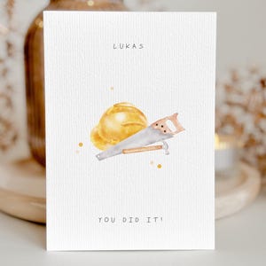 Könnte beinhalten: Weiße Karte mit dem Namen "LUKAS" oben und dem Satz "YOU DID IT!" unten. Die Karte zeigt eine Aquarellillustration eines gelben Bauhelms und einer Handsäge.