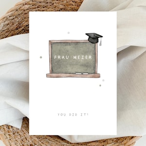 Könnte beinhalten: Eine Abschlusskarte mit einem Tafeldessign. Die Tafel zeigt den Text "FRAU MEIER" und eine Abschlusskappe. Die Karte enthält auch den Text "YOU DID IT!" in einer einfachen Schriftart. Die Karte ist vor einem geflochtenen Korb und einem weißen Tuch platziert.