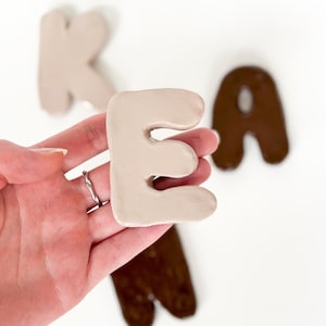 Puede incluir: Mano sosteniendo una letra "E" de cerámica beige con otras letras en el fondo. Las letras están en una fuente redondeada. Las otras letras son una "K" beige y una "A" marrón. El fondo es blanco.