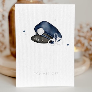 Op de afbeelding: Een wenskaart met een illustratie van een marineblauwe politiepet en handboeien. De pet heeft een zwarte klep met de naam "CHRIS" erop gedrukt. De tekst "YOU DID IT!" staat onderaan de kaart.