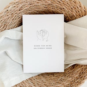 May include: A white card with a line drawing of two hands holding a heart. The text on the card reads "Danke f&uuml;r deine haltenden H&auml;nde".