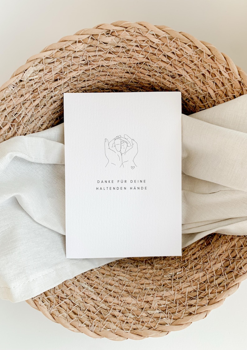 May include: A white card with a line drawing of two hands holding a heart. The text on the card reads "Danke f&uuml;r deine haltenden H&auml;nde".