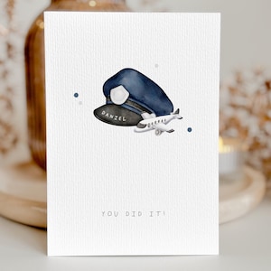 May include: Greeting card with a watercolor illustration of a pilot's hat and a small airplane. The hat is navy blue with a black brim that reads "DANIEL". The text "YOU DID IT!" is at the bottom.