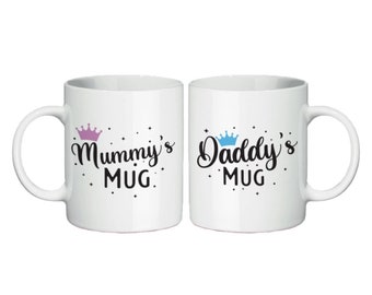 May include: Two white ceramic mugs with black text and crown designs. The mug on the left says "Mummy's Mug" with a pink crown. The mug on the right says "Daddy's Mug" with a blue crown.