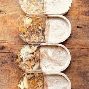 Può includere: Quattro vassoi ovali in resina con un designo marmoreo bianco e beige. Ogni vassoio ha una metà trasparente riempita di fiori e piante secche.