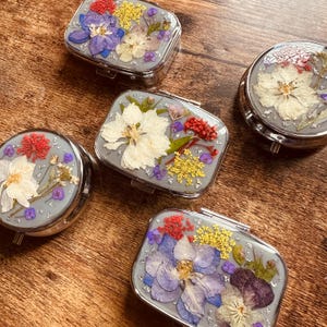 Peut inclure: Cinq petites boîtes à pilules argentées avec différents motifs de fleurs pressées. Les boîtes sont rectangulaires et rondes, avec un couvercle argenté et une base transparente. Les fleurs sont pressées entre le couvercle et la base, créant un design magnifique et unique.