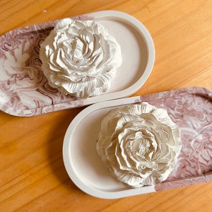May include: Two white ceramic flower-shaped decorative objects on marble-patterned oval trays. The trays are white with brown and pink marbling.
