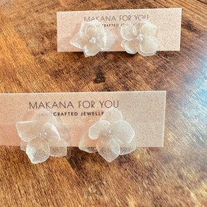 Könnte beinhalten: Ein Paar zarte weiße Blumenohrringe mit Perlenakzenten. Die Ohrringe werden auf einer Karte mit dem Text "Makana For You Crafted Jewell" präsentiert.