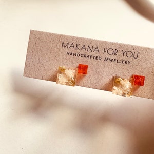 Könnte beinhalten: Ein Paar quadratische Ohrringe aus Harz mit goldenen Flocken und einem roten Akzent. Die Ohrringe werden auf einer Karte mit der Aufschrift "Makana For You Handcrafted Jewellery" präsentiert.