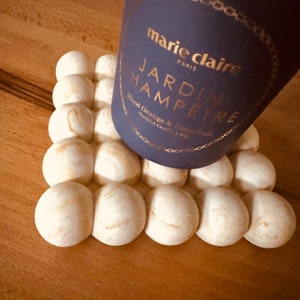 May include: A purple candle jar with gold lettering that reads "marie claire PARIS JARDIN HAMPTRE Blood Orange & Grapefruit Scented Candle 170g" sitting on a white wooden coaster.