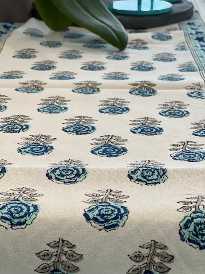 May include: White table runner with a repeating pattern of blue and green floral designs. The flowers are stylized and have a vintage look.