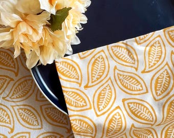 May include: A white fabric placemat with a yellow leaf pattern. The placemat is folded in half and placed on top of a black plate. A bouquet of yellow flowers is placed on the plate.