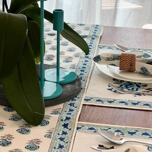May include: A white table runner with a blue and green floral pattern. The runner has a border of blue stars and is placed on a wooden table. Two teal candle holders are on the table in the background.