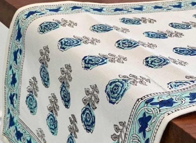 May include: White table runner with a blue and green floral pattern. The runner has a border with a repeating design of blue and green leaves and flowers.