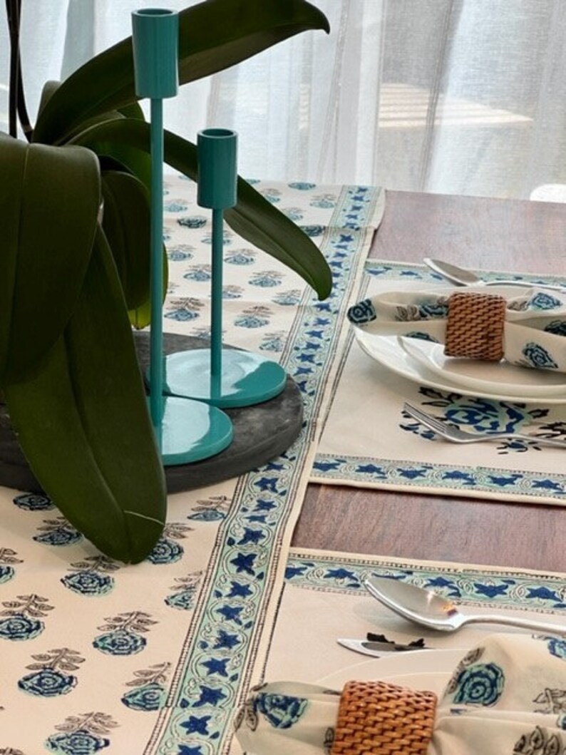 May include: A white table runner with a blue and green floral pattern. The runner has a border of blue stars and is placed on a wooden table. Two teal candle holders are on the table in the background.